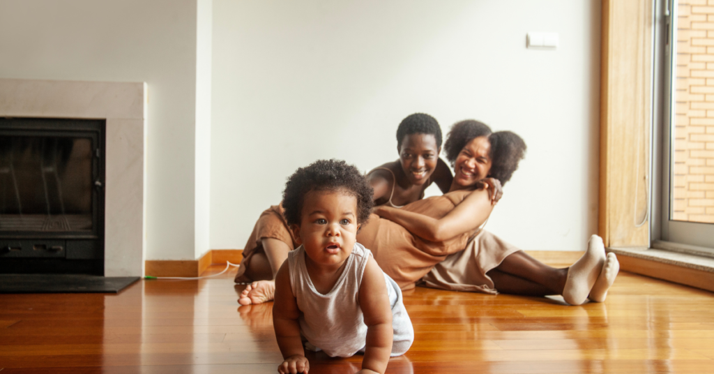 black lesbian couple embracing on the floor with their baby crawling in front of them
