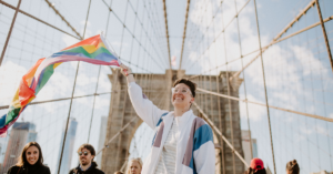 masc presenting queer person waving a Pride flag on the Brooklyn Bridge