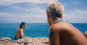 A dreamy seaside scene from Ripe! showing two young women in swimsuits sitting on rocky cliffs beside bright blue ocean water, sharing a quiet, emotionally charged moment.